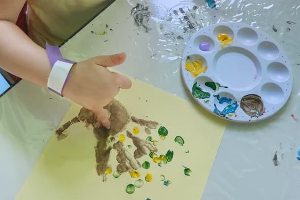 Mão de criança coberta de tinta ao lado da paleta de cores, em oficina de arte